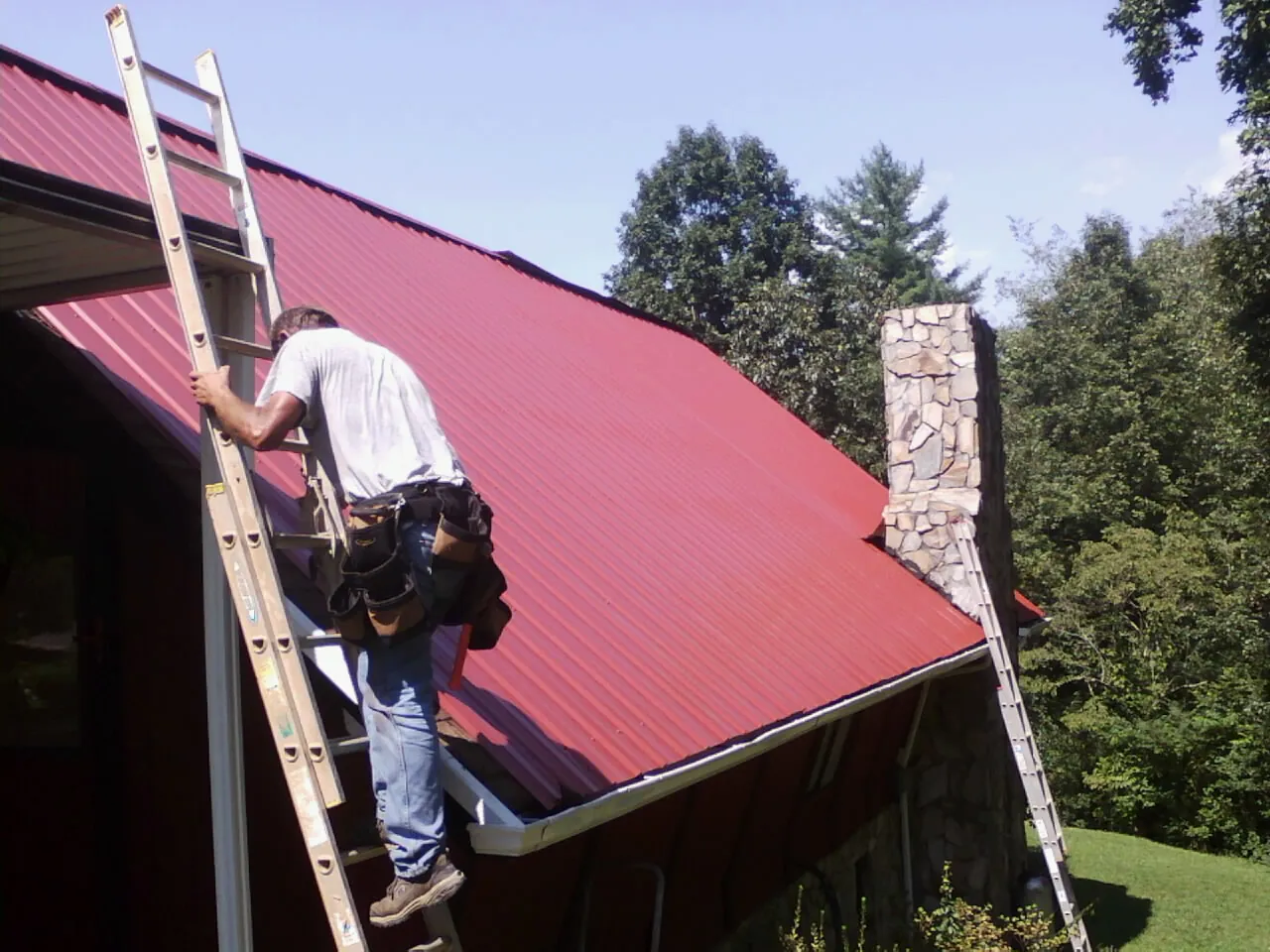 Roofer on a ladder beside a red metal roof and stone chimney — Soffit & Fascia Repair by Roofing Brothers in Fields